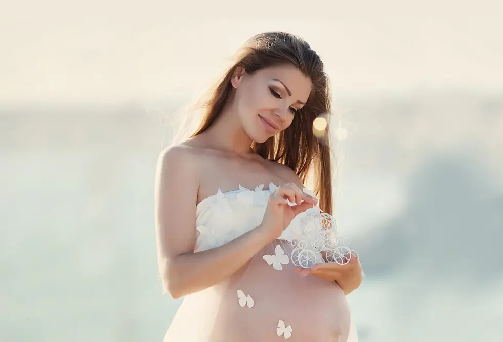 Beautiful pregnant woman in rich pink dress holds hands on her belly posing in luxury white room беременность без проблем